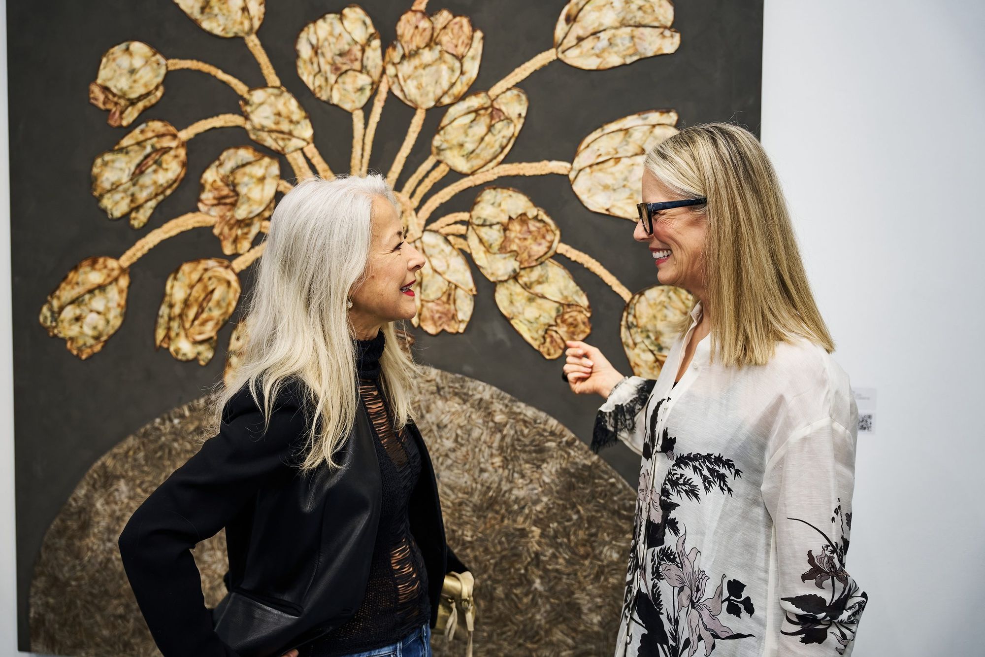 Two gallery visitors in conversation in front of a large painting of pale gold flowers on a dark ground