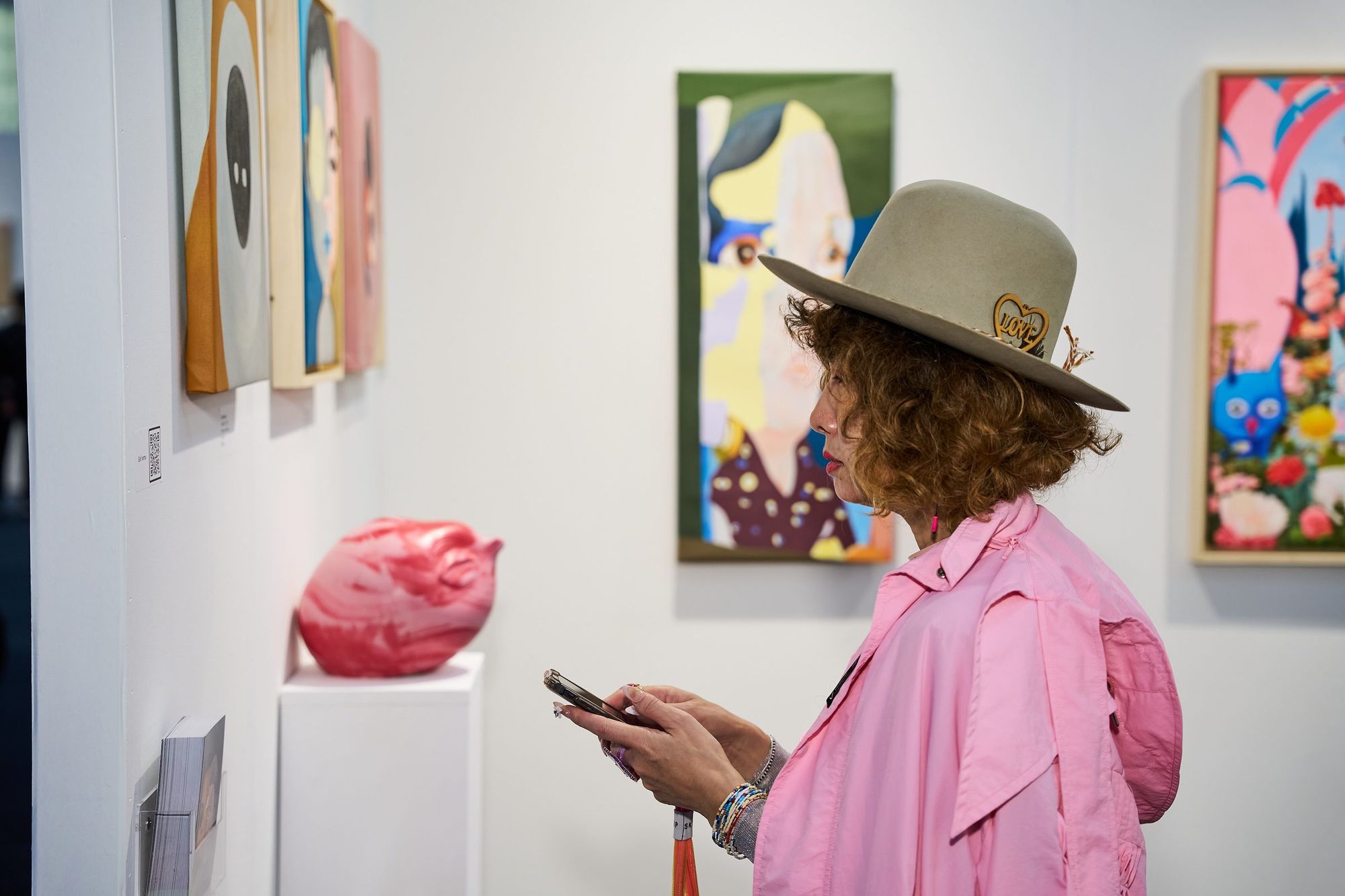 A visitor in a pink coat and felt hat pauses at a wall of framed paintings beside a pink sculpture on a plinth
