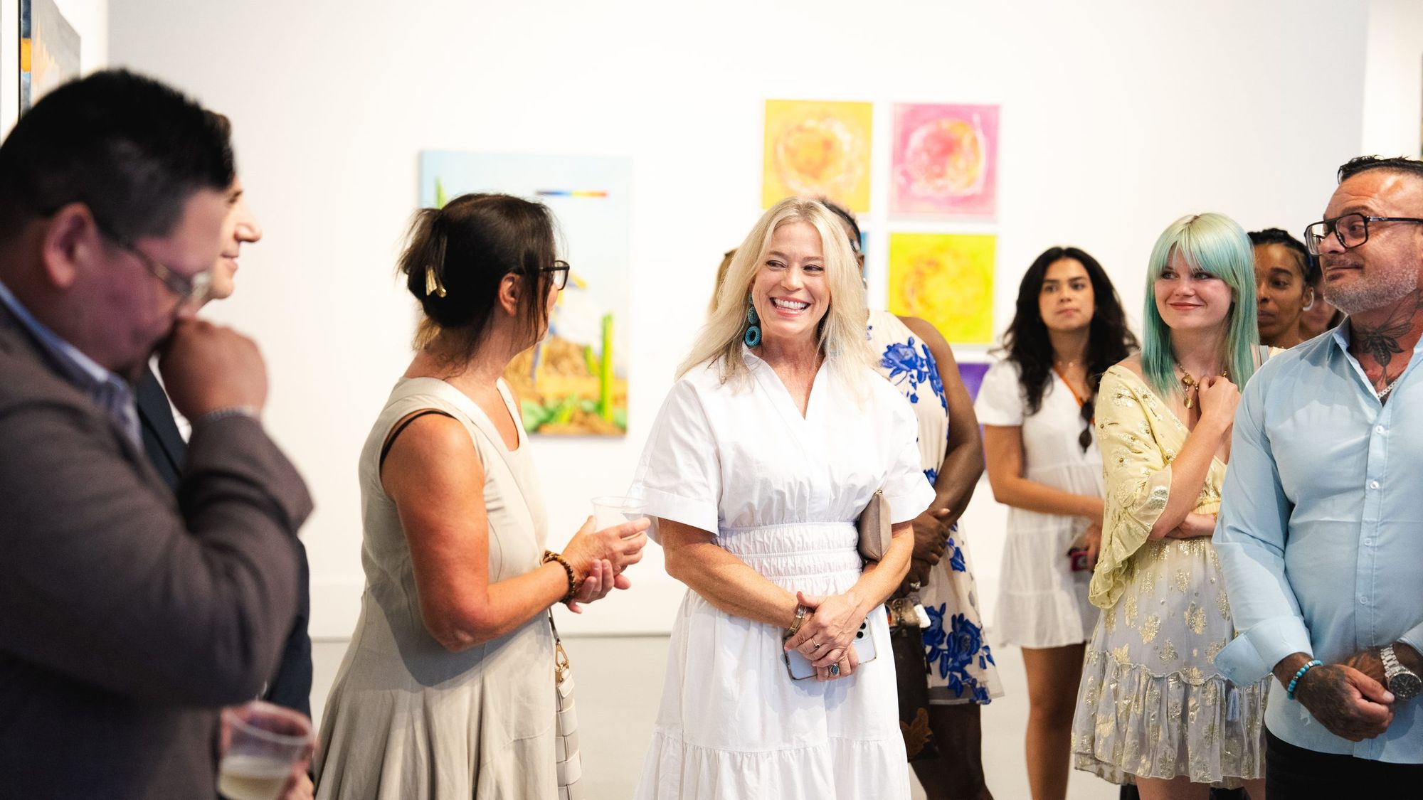 Guests with drinks talking at an exhibition opening, colourful works on the walls behind them