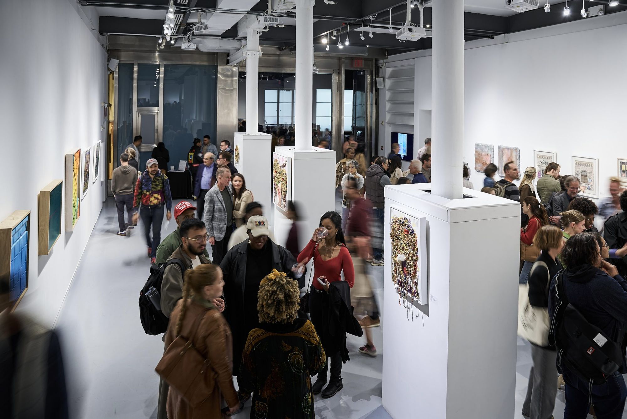 A crowded art fair floor seen from above, visitors moving between white plinths and walls of framed works