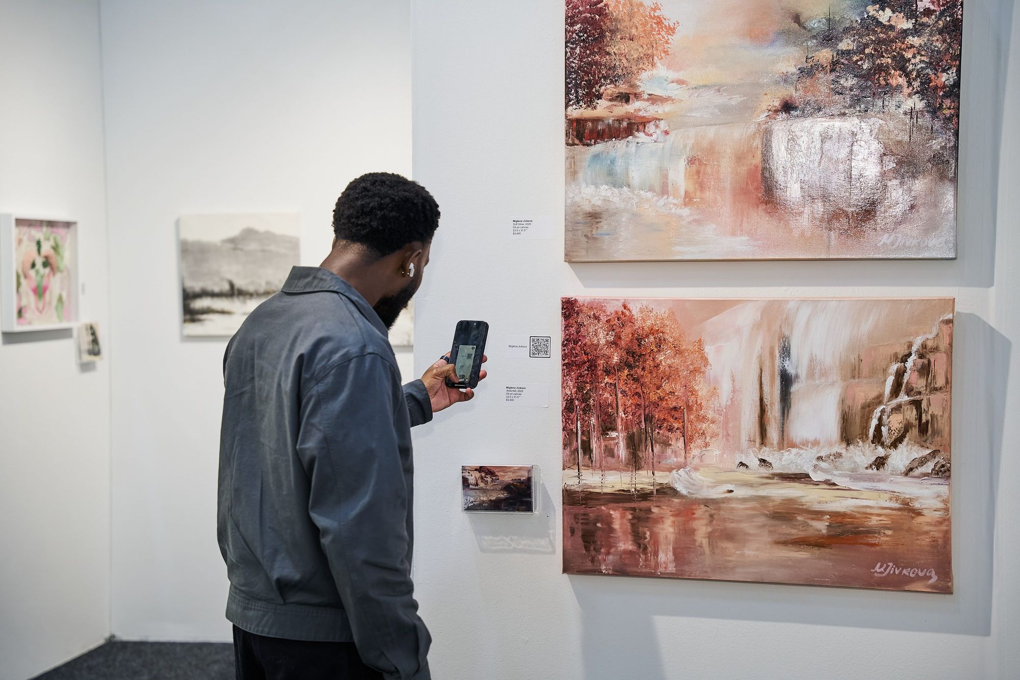 A visitor photographing two framed landscape paintings of autumn trees and a waterfall with his phone